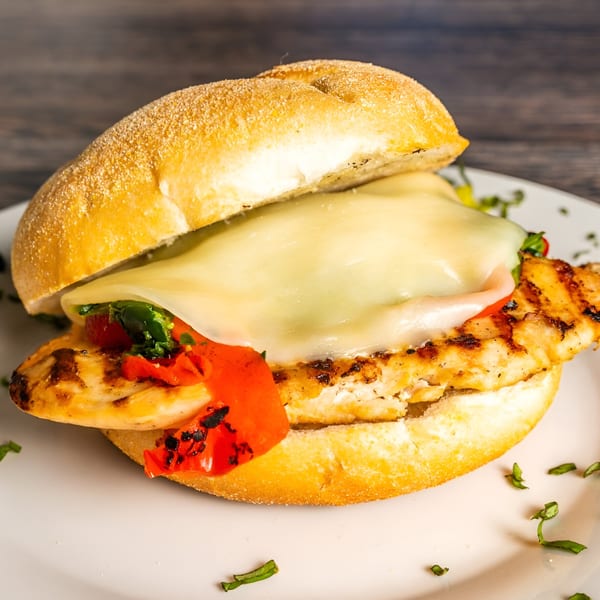 Grilled chicken sandwich with melted cheese, roasted red peppers, and greens on a bun.