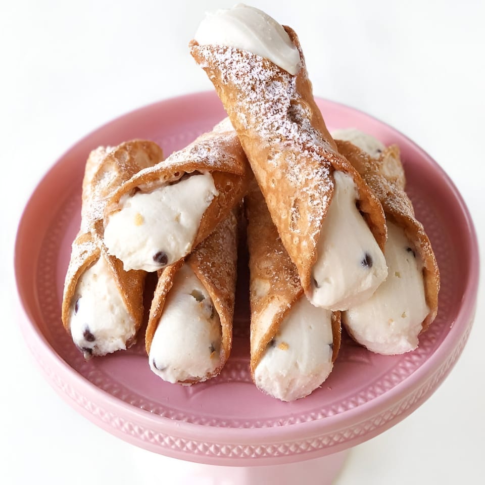 A pink cake stand holds several cannoli, filled with cream and chocolate chips, dusted with powdered sugar.